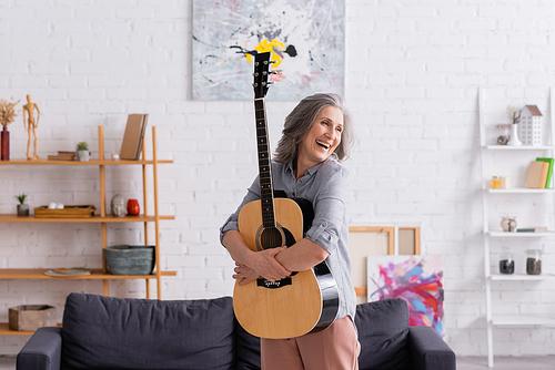 cheerful mature woman with grey hair embracing acoustic guitar near couch in living room
