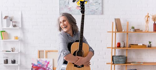 joyful mature woman with grey hair standing with acoustic guitar in living room, banner