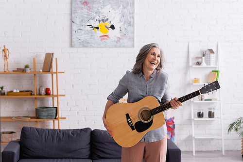 happy mature woman with grey hair standing with acoustic guitar and laughing in living room