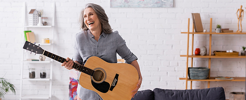 happy mature woman with grey hair standing with acoustic guitar in living room, banner
