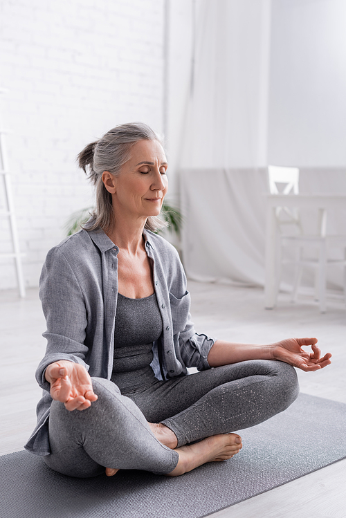 mature woman with grey hair sitting in lotus pose while meditating on yoga mat