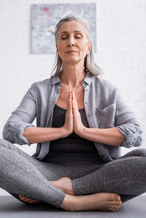 mature woman with praying hands and closed eyes sitting in lotus pose on yoga mat
