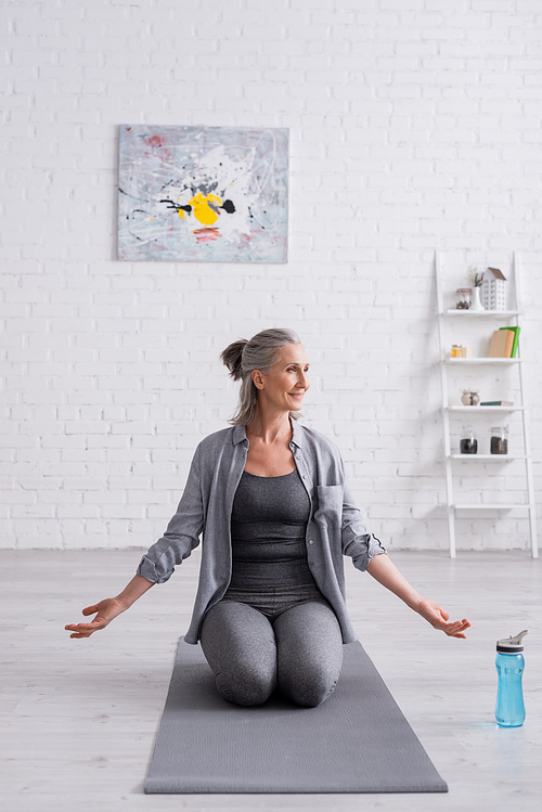 pleased mature woman with grey hair practicing yoga near sports bottle