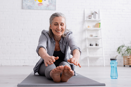 happy mature woman with grey hair stretching on yoga mat near sports bottle