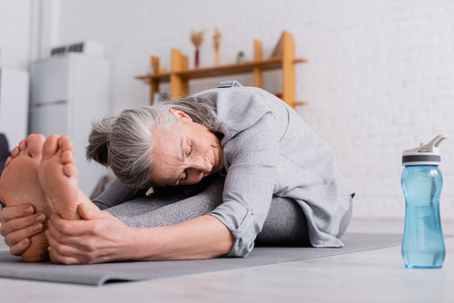 middle aged woman with grey hair stretching on yoga mat near sports bottle