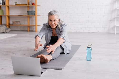 mature woman with grey hair stretching on yoga mat near sports bottle and laptop