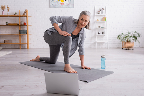 mature woman with grey hair practicing on yoga mat near sports bottle while watching tutorial on laptop