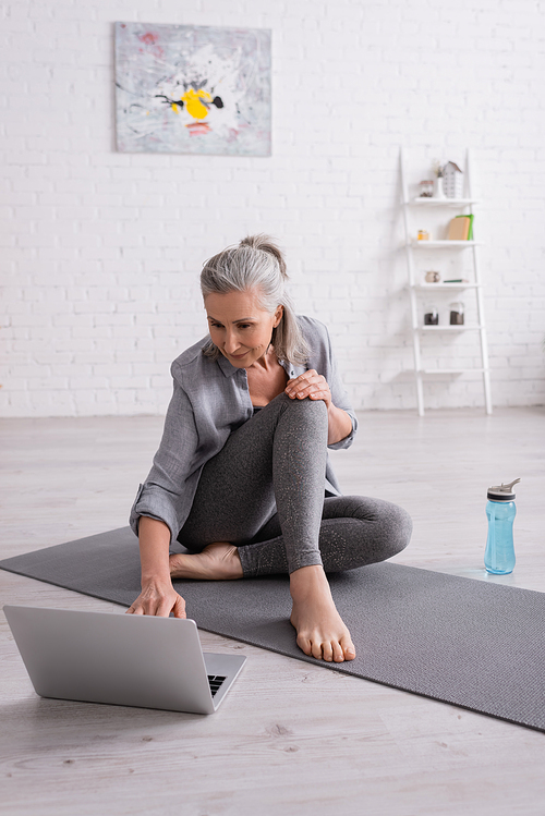 mature woman with grey hair sitting on yoga mat while watching tutorial on laptop