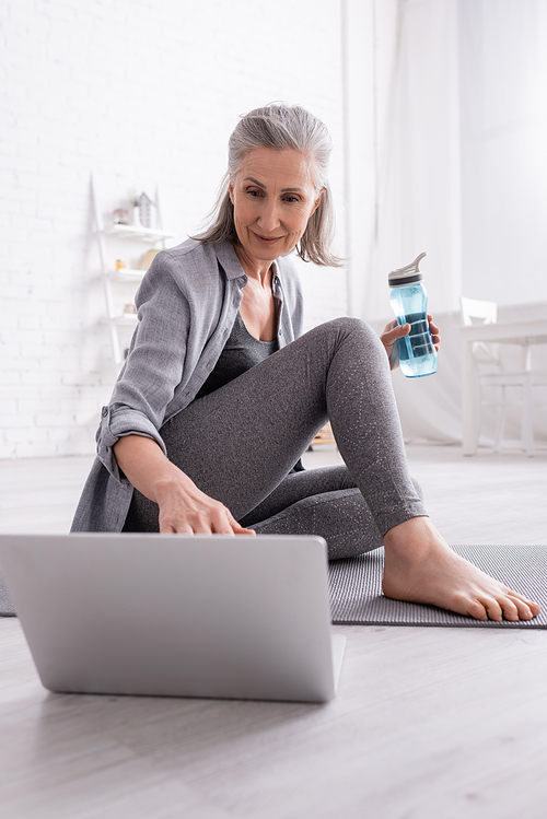 mature woman with grey hair sitting on yoga mat while watching tutorial on laptop and holding sports bottle