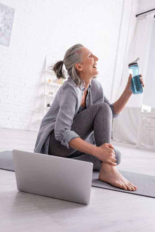 happy mature woman with grey hair sitting on yoga mat near laptop and holding sports bottle