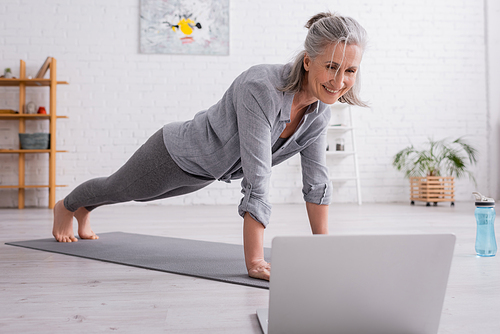 happy mature woman practicing and watching yoga tutorial on blurred laptop