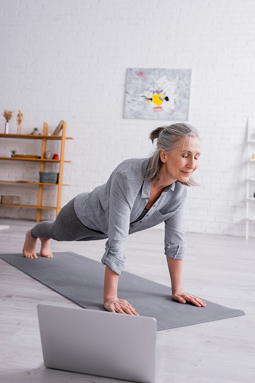 mature woman doing plank on fitness mat near laptop