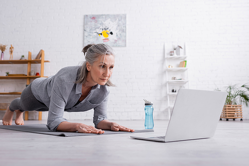 middle aged woman doing plank on fitness mat near laptop