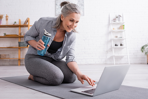 smiling middle aged woman sitting on fitness mat, holding sports bottle and using laptop