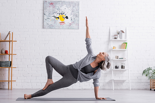 flexible middle aged woman in sportwear with raised hand practicing yoga on mat at home