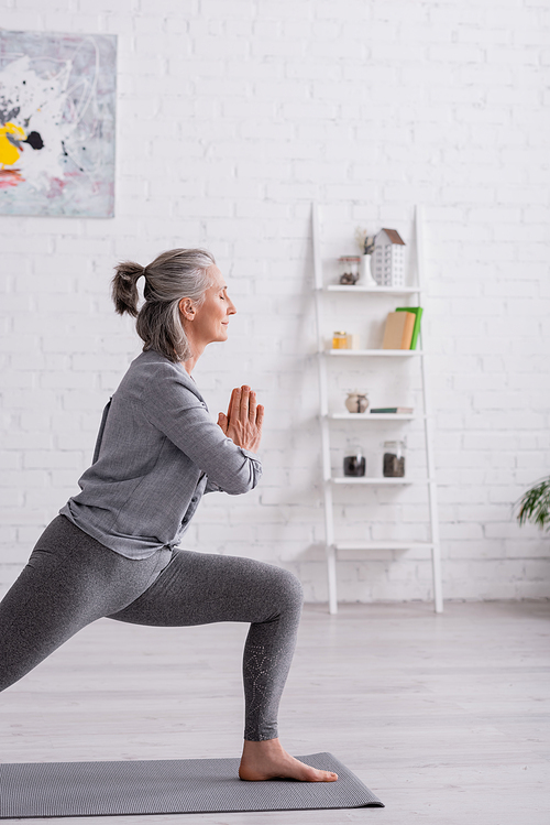 middle aged woman in warrior with praying hands pose practicing yoga on mat at home