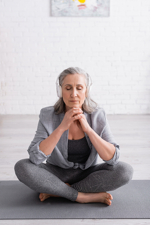 middle aged woman in wireless headphones meditating while sitting in lotus pose on yoga mat
