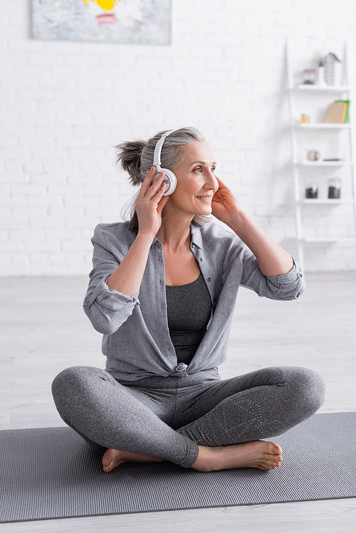 happy middle aged woman in wireless headphones sitting in lotus pose on yoga mat at home