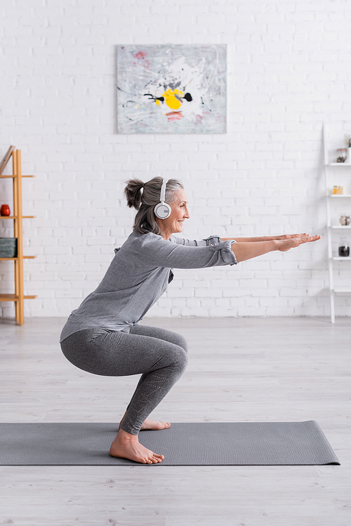 side view of cheerful middle aged woman in sportwear and headphones doing squat on mat at home