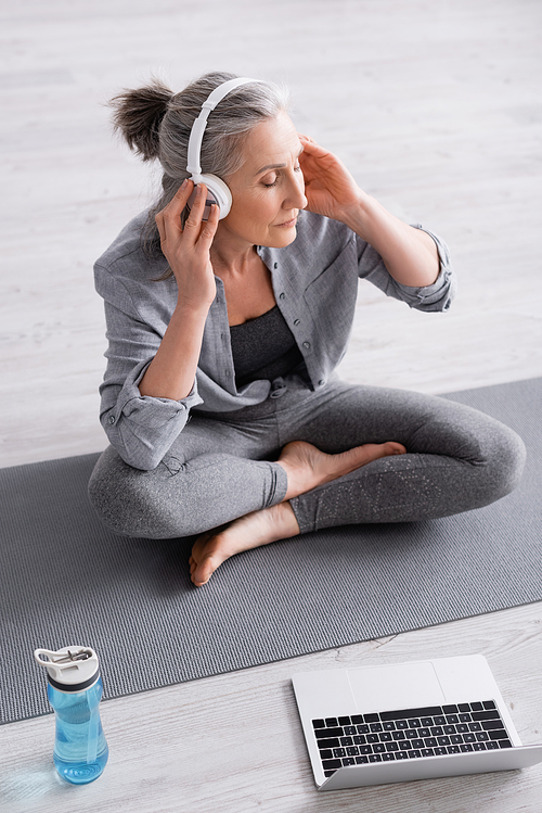 high angle view of middle aged woman in wireless headphones meditating while sitting in lotus pose on yoga mat near laptop