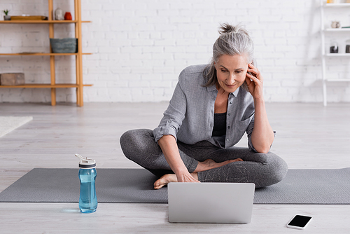 middle aged woman in sportwear sitting in lotus pose on yoga mat and watching online tutorial on laptop