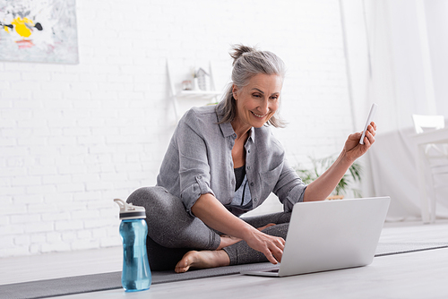 happy middle aged woman sitting in lotus pose on yoga mat and watching online tutorial on laptop and holding smartphone
