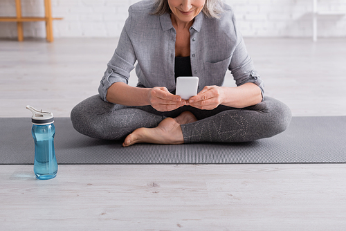 cropped view of middle aged woman sitting in lotus pose on yoga mat and using smartphone