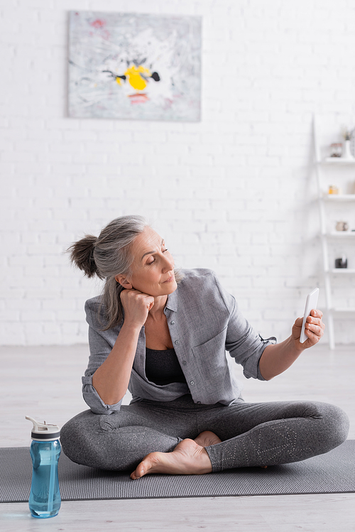 middle aged woman sitting in lotus pose on yoga mat and using smartphone