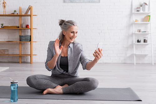 middle aged woman sitting in lotus pose on yoga mat and waving hand while having video call on smartphone