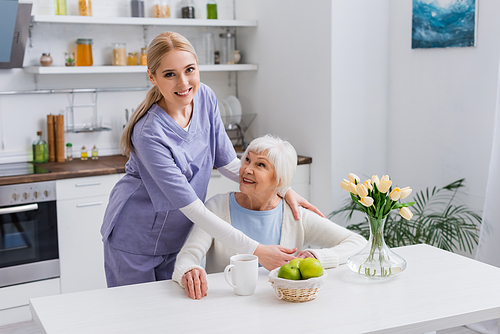 young nurse smiling at camera while embracing happy aged woman in kitchen