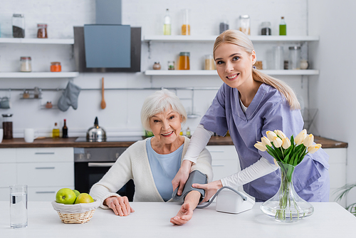 happy social worker and aged woman  while measuring blood pressure in kitchen