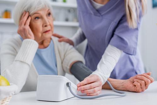aged woman touching head while nurse measuring her blood pressure with tonometer, blurred background