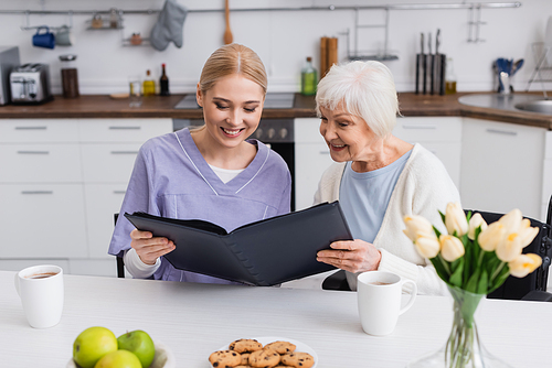 pleased senior woman and nurse looking at photo album in kitchen