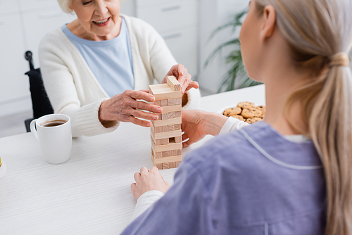 cropped view of joyful senior woman playing wood blocks game with nurse on blurred foreground