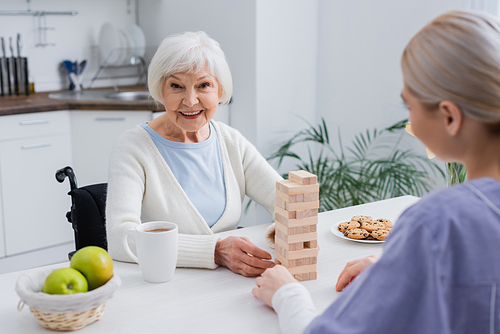 aged disabled woman smiling at camera while playing wood blocks game with nurse