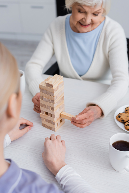 cheerful elderly woman playing wood blocks game with nurse on blurred foreground