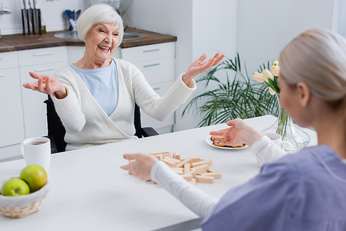 excited senior woman showing wow gesture near nurse pointing at wooden blocks on table