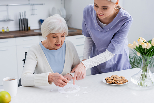 young nurse pointing at jigsaw puzzle near elderly woman in kitchen