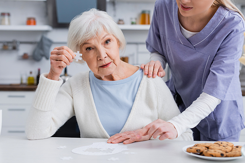elderly woman showing piece of jigsaw puzzle while nurse touching her shoulder