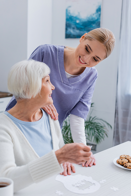 smiling nurse looking at happy aged woman playing jigsaw puzzle at home