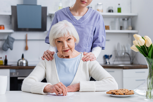 young nurse touching shoulders of elderly woman  near jigsaw puzzle