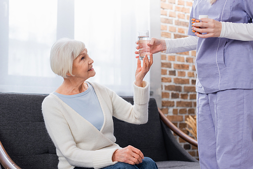 social worker holding pills while giving glass of water to smiling elderly woman