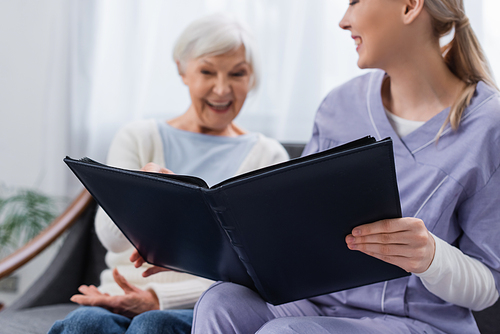 cheerful senior woman with social worker looking at photo album at home, blurred background