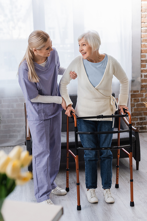 young nurse supporting smiling senior woman walking in living room with medical walkers