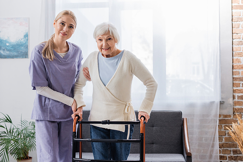 happy nurse and senior woman  near medical walkers in living room