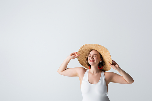 overweight and pleased woman in earrings and swimsuit adjusting straw hat isolated on white