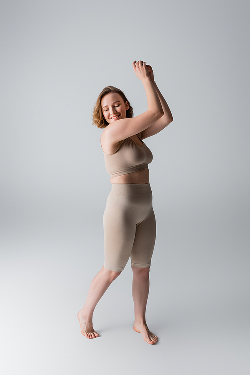 full length of overweight and barefoot woman in lingerie smiling while posing on grey