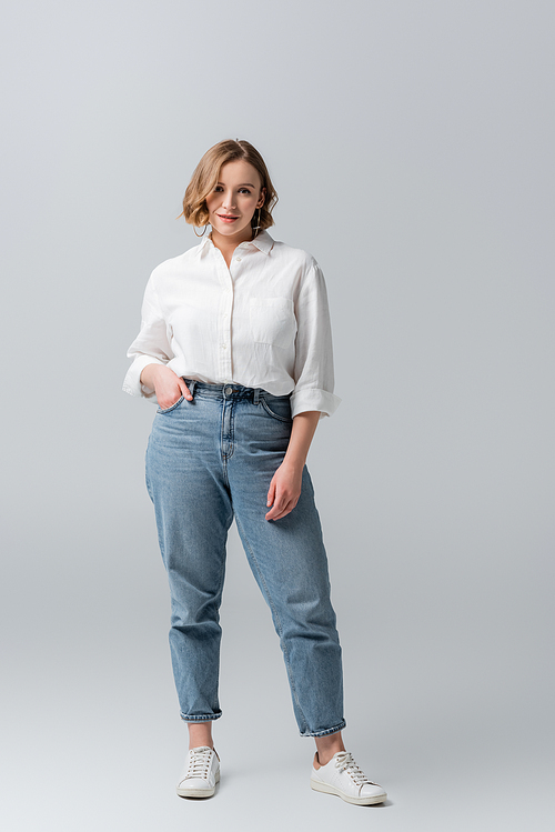 full length of overweight young woman in jeans posing with hand in pocket on grey