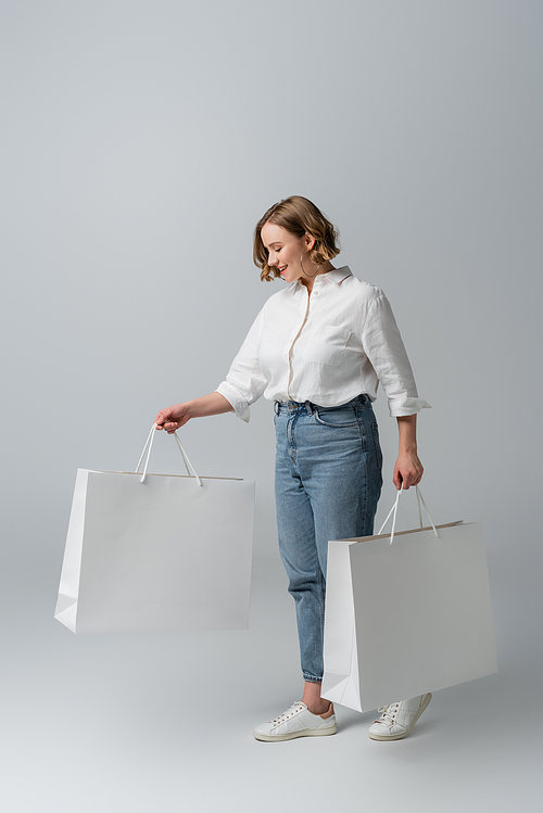 happy overweight woman in jeans and white shirt holding shopping bags on grey