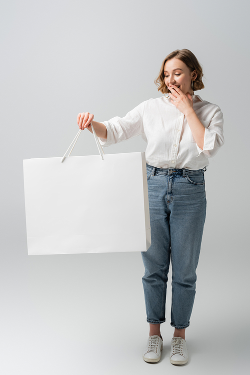 full length of excited overweight woman in jeans and white shirt holding shopping bag on grey